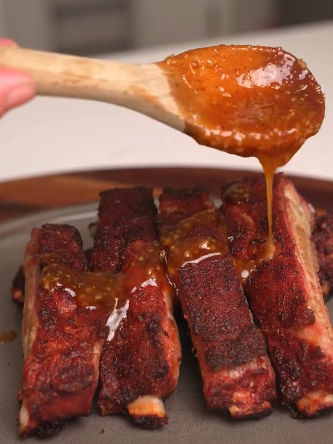 A wooden spoon drizzles a golden-brown sauce over a plate of spiced, cooked pork ribs arranged in neat rows. The ribs have a dark, savory crust and sit on a gray plate.