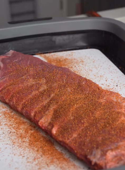 A slab of raw pork ribs is covered with a reddish-brown dry spice rub, resting on a white surface inside a black tray, ready for cooking or grilling.