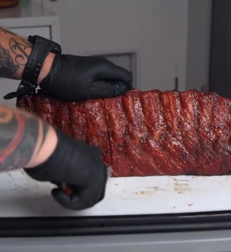 A person wearing black gloves and a smartwatch slices a rack of barbecued ribs on a white cutting board. The ribs have a dark, glossy, seasoned crust.