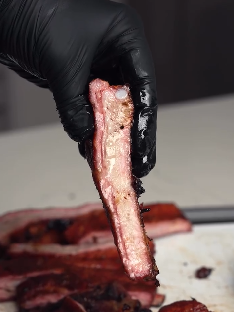 A hand wearing a black glove holds up a single, juicy pork rib with a smoky, caramelized crust. More ribs are visible, sliced on a cutting board in the background.