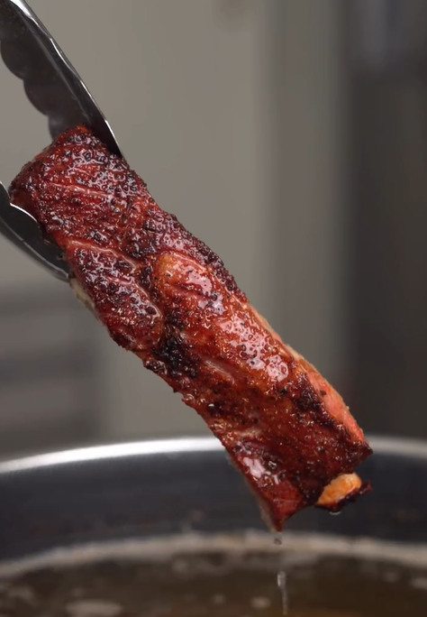 A close-up of metal tongs holding a crispy, seasoned strip of bacon above a pan, with oil dripping from the bacon.