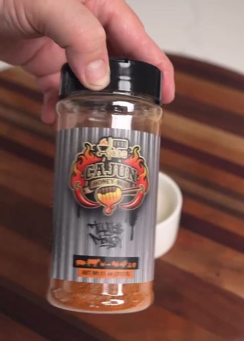 A hand holding a bottle of Meat Church BBQ Cajun Honey Seasoning over a wooden surface, with a small white bowl in the background.