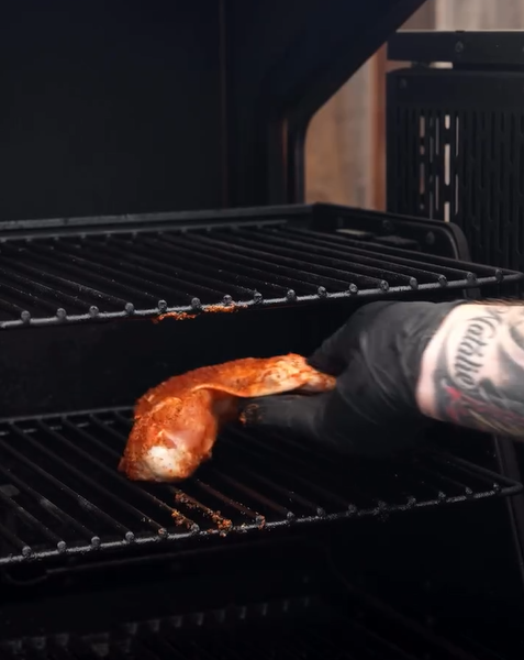 A tattooed person wearing a black glove places a seasoned piece of meat onto the rack of a smoker or grill.