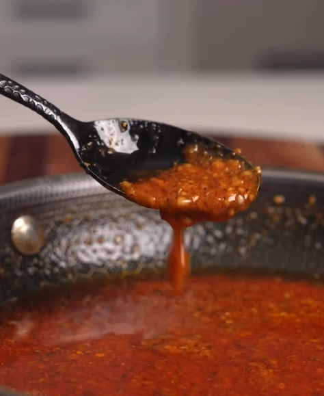 A close-up of a spoon lifting thick, red pasta sauce from a pan, with sauce dripping off the spoon.