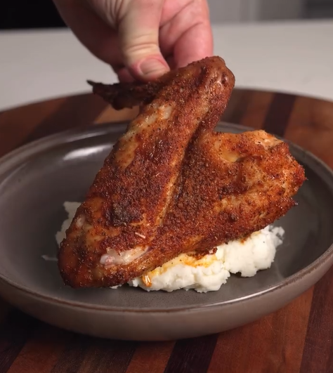 A hand places a seasoned, cooked chicken wing on a mound of mashed potatoes on a gray plate, which sits on a wooden surface.