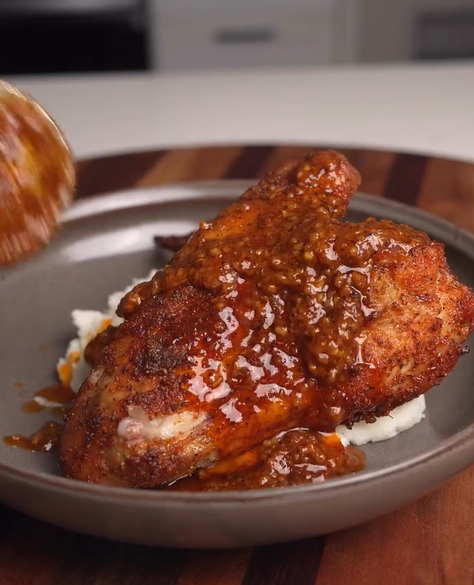 A cooked chicken breast topped with spicy brown sauce sits on a bed of mashed potatoes on a gray plate, resting on a wooden surface. A spoon is shown pouring extra sauce onto the chicken.