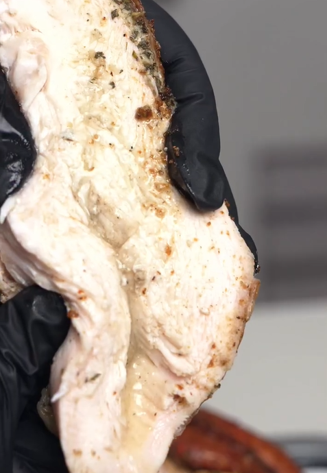 A person wearing a black glove holds up a thick, juicy slice of cooked chicken breast seasoned with herbs. The background is out of focus.