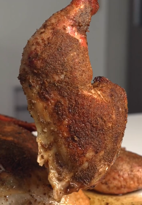 A close-up of a seasoned and cooked chicken leg quarter with crispy skin, displaying a rich blend of spices and visible texture, held upright against a blurred indoor background.
