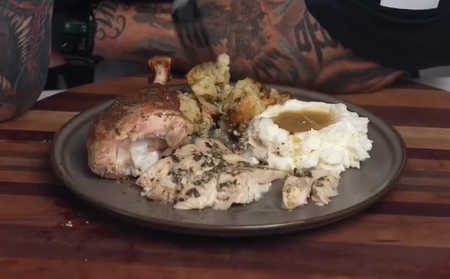 A plate of food with roasted chicken, mashed potatoes with gravy, roasted cauliflower, and sliced herb-seasoned chicken on a wooden table with tattooed arms visible in the background.