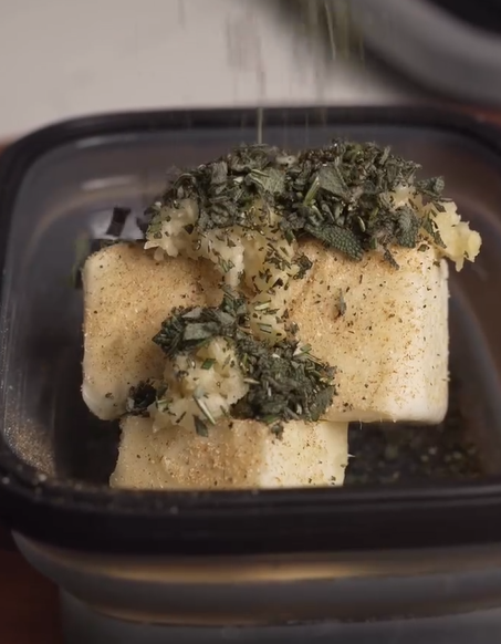 Two blocks of butter in a black container are topped with minced garlic, chopped herbs, and spices, with more herbs being sprinkled from above.