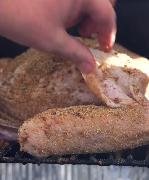 A close-up of a hand lifting the skin of a raw, seasoned turkey to apply spices underneath while it rests on a grill grate.