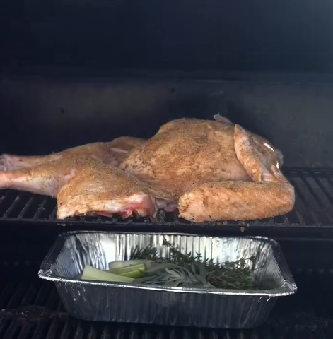 A seasoned, spatchcocked chicken is grilling on a rack, with a foil tray of herbs and vegetables, including celery and rosemary, placed below it inside the grill.