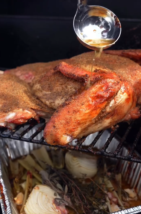 A seasoned, roasted chicken is being basted with juices from a ladle. Below the chicken, onions, herbs, and other aromatics are visible in a foil pan, likely to add flavor and catch drippings.