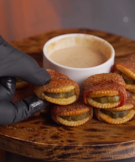 A gloved hand picks up a crispy snack made of crackers, pickles, and seasoning, stacked near a bowl of dipping sauce on a wooden serving board.