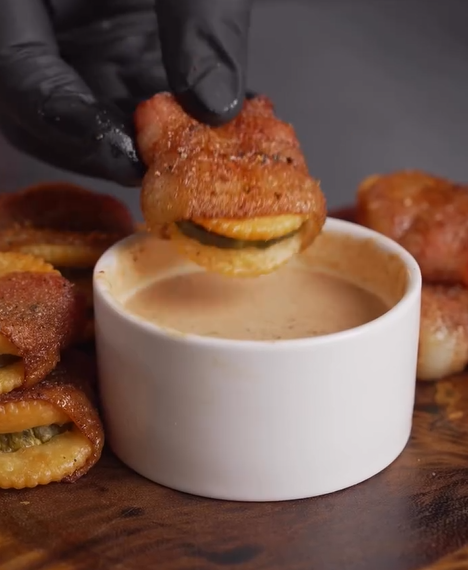 A hand in a black glove dips a crispy, golden-brown stuffed ravioli into a creamy dipping sauce in a white bowl. More stuffed ravioli pieces are arranged on a wooden surface nearby.