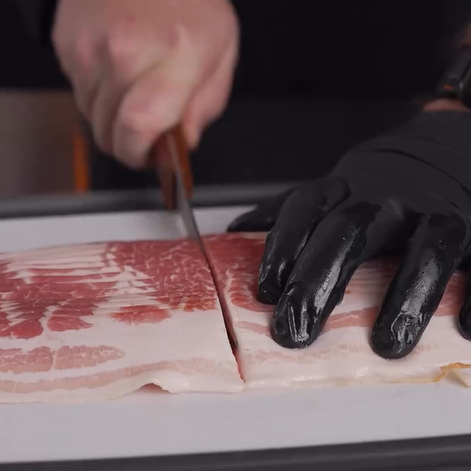 A person wearing a black glove slices a thick piece of raw bacon on a cutting board with a large knife.