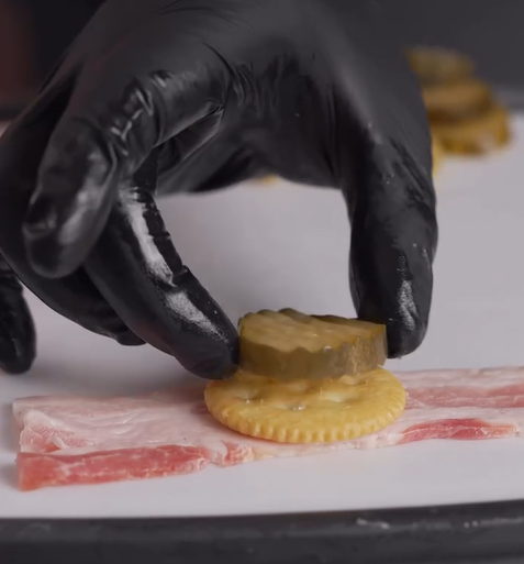 A hand in a black glove places a pickle slice onto a round cracker, which is resting on a strip of raw bacon on a white surface.
