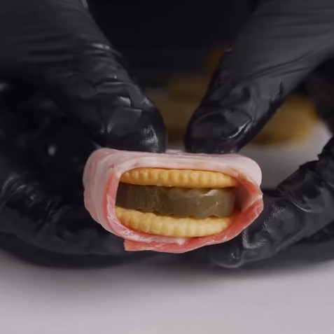 A person wearing black gloves holds a sandwich made of two round crackers with a pickle slice in the middle, all wrapped in a strip of raw bacon.