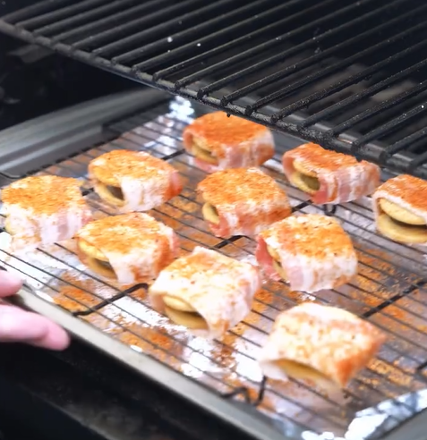 Several bacon-wrapped, seasoned food items are arranged on a wire rack inside a grill, ready to be cooked. A hand is visible on the left, holding the rack in place.