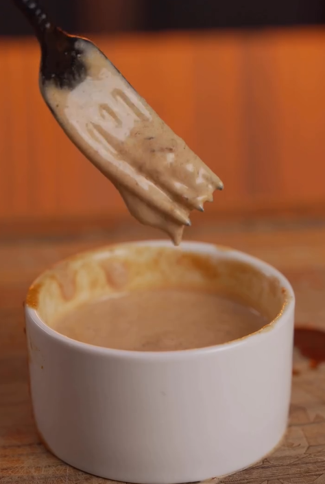 A close-up of a fork holding a thick, creamy brown sauce above a white ramekin filled with the same sauce. The background is blurred with warm orange tones.