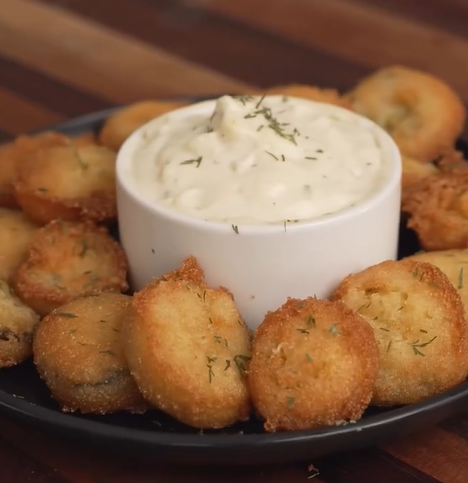A plate of golden-brown fried mushrooms arranged around a bowl of creamy dipping sauce, garnished with herbs, on a wooden surface.