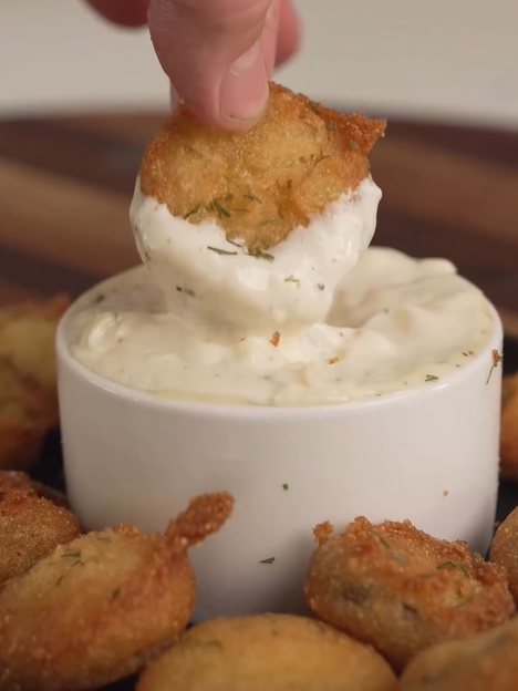 A hand dips a crispy, breaded fried pickle slice into a bowl of creamy white ranch dipping sauce, with more fried pickles arranged around the bowl.