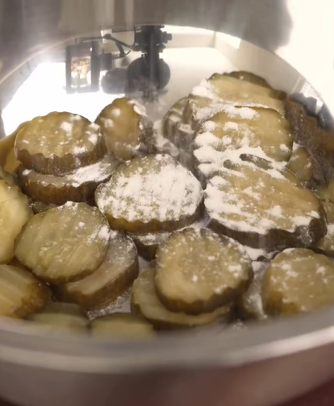 Sliced pickles in a metal bowl being dusted with white powder, possibly a seasoning or salt, under a small automated dispenser.