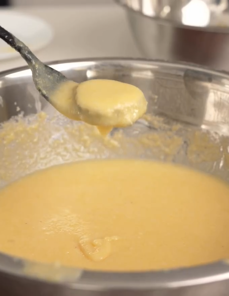 A spoon holds up thick, yellow cake batter over a stainless steel mixing bowl filled with the same batter. Another empty bowl is visible in the background.