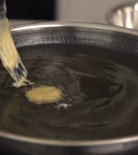 A close-up of batter being dropped into hot oil in a pan, starting to fry and form a round fritter.