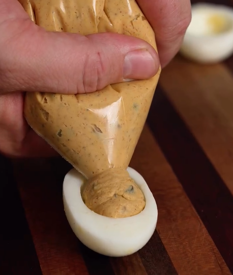 A hand squeezes a piping bag filled with spiced filling into a halved hard-boiled egg on a wooden surface, making deviled eggs.