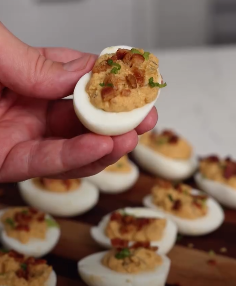 A hand holds a deviled egg topped with chopped bacon and herbs. More deviled eggs are arranged on a wooden board in the background.