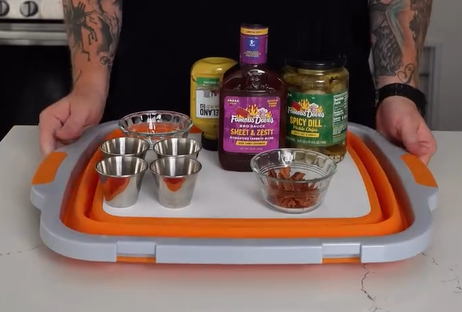 A person holds a tray with metal cups, bowls of spices and pickles, a bottle of barbecue sauce, and a bottle of mustard on a white and orange cutting board.