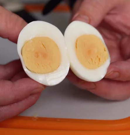 Two hands hold a hard-boiled egg sliced in half lengthwise, revealing a firm, pale yellow yolk and a smooth white exterior. The egg rests above an orange-edged cutting board.