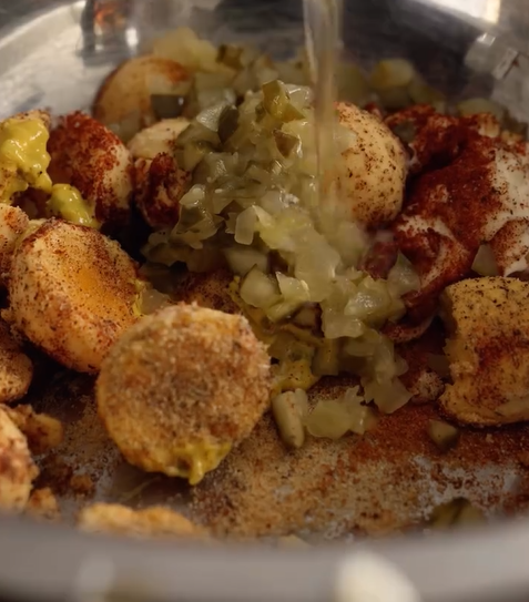A close-up of boiled egg yolks, chopped pickles, and seasonings in a metal mixing bowl, with liquid being poured in, likely in preparation for making deviled eggs.