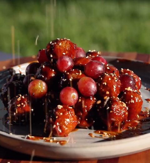 A plate of crispy, sauce-covered chicken bites topped with fresh red grapes, sesame seeds, and a sticky glaze drizzle, set outdoors on a wooden table with blurred green grass in the background.