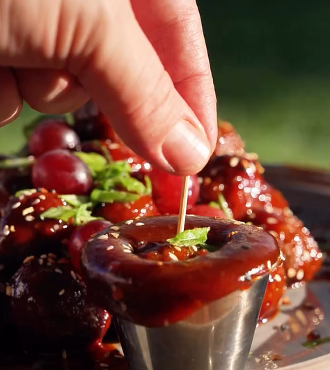A hand dips a toothpick into a metal cup filled with dark, glossy sauce, garnished with a small green herb, with barbecue-glazed meatballs and grapes in the background.