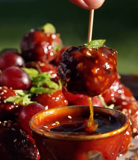 A hand dips a glossy, sauce-covered round fruit on a skewer into a small bowl of sauce. In the background, similar coated fruits are garnished with sesame seeds and mint leaves.
