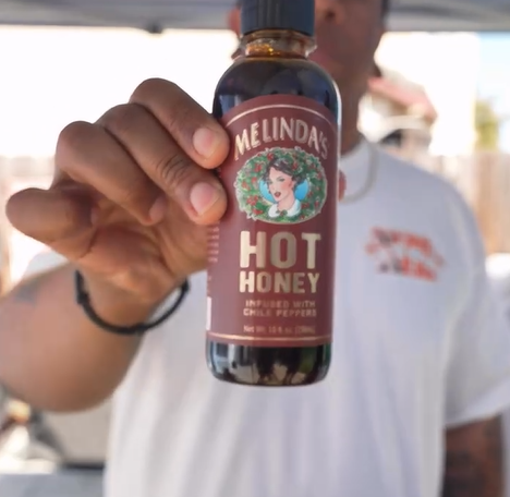 A person holds up a bottle of Melinda’s Hot Honey infused with chile peppers, with the label and logo clearly visible. The background is blurred.