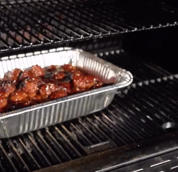 A disposable aluminum tray filled with glazed, cooked meat sits on a grill rack inside a barbecue smoker or grill.