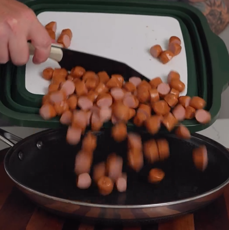 A hand uses a spatula to transfer sliced sausages from a cutting board into a black frying pan on a wooden countertop.