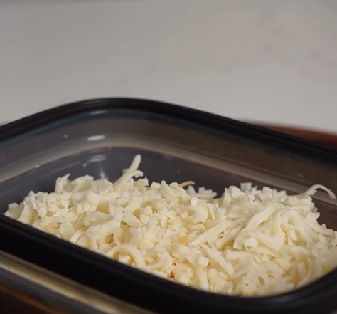 A close-up of shredded cheese in a rectangular black plastic container on a light-colored surface.