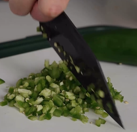 A close-up of a hand using a black knife to finely chop green peppers on a white cutting board.