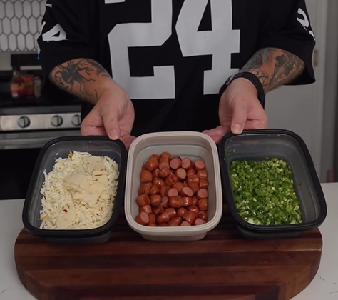 A person holding three containers: one with grated cheese, one with sliced sausages, and one with chopped green peppers, on a wooden cutting board.