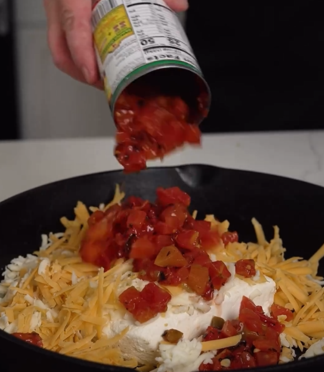 A hand pours canned diced tomatoes and green chilies over a mixture of shredded cheese and a block of cream cheese in a black skillet.