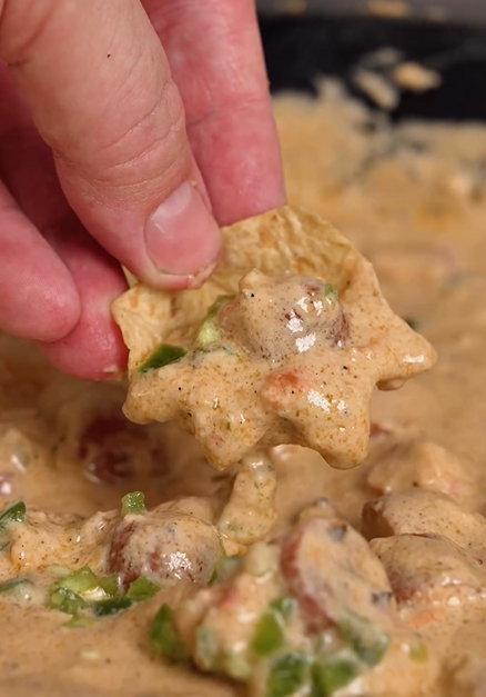 A hand dips a tortilla chip into creamy queso dip with pieces of sausage and chopped green peppers visible in the mixture.