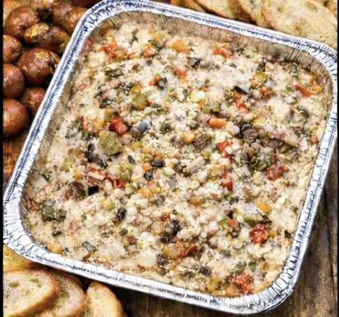A foil tray filled with a colorful, chunky vegetable mixture, likely a baked casserole, surrounded by slices of toasted baguette and roasted whole mushrooms on a wooden surface.