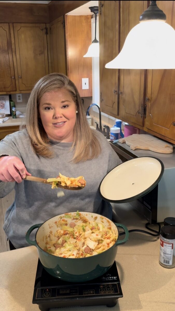 A woman in a gray sweater stands in a kitchen, holding a spoonful of food above a pot of stew on the stove. She smiles at the camera while holding the pot lid in her other hand. Wooden cabinets and a lamp are in the background.