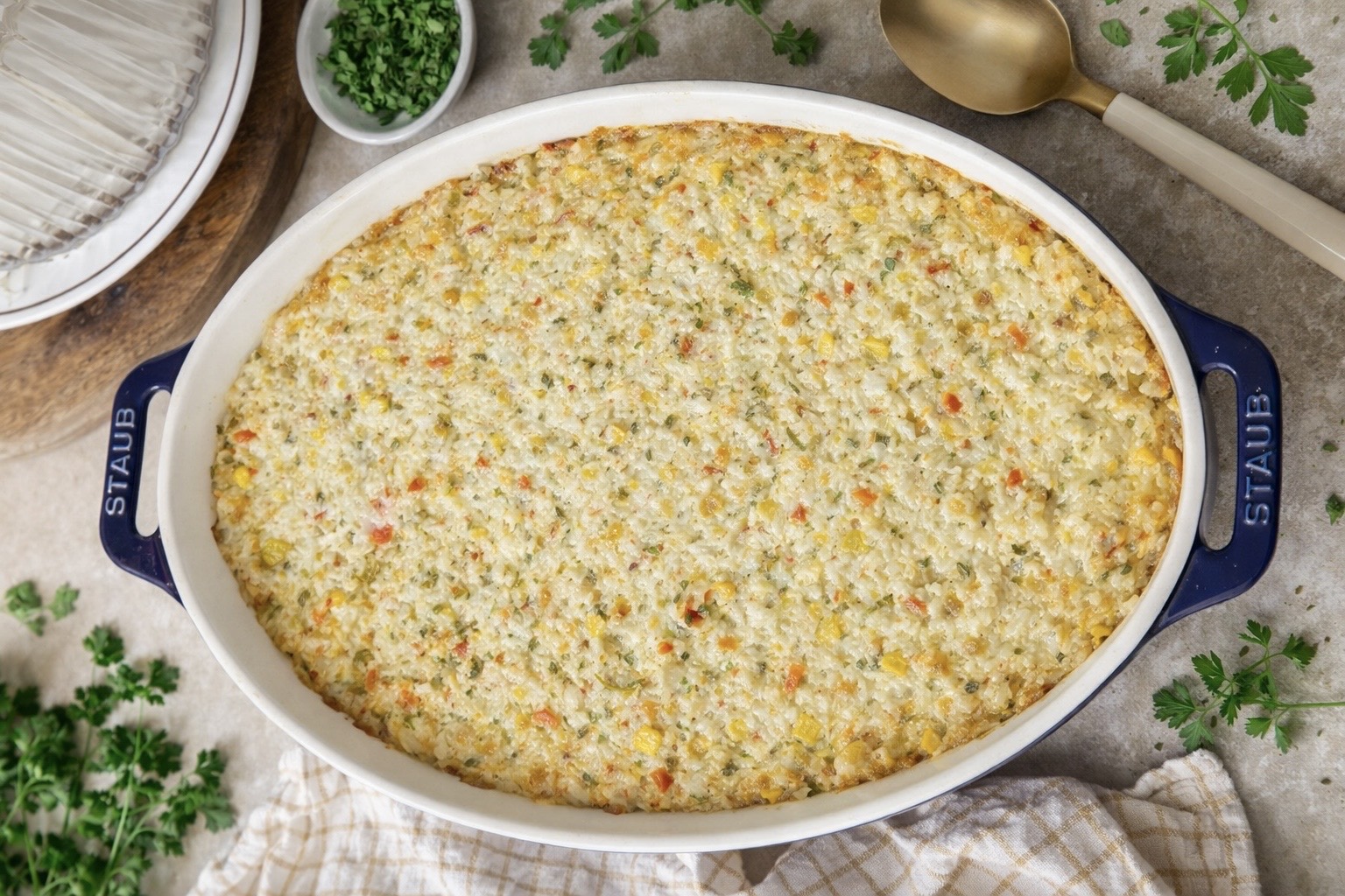 An oval casserole dish filled with a golden-baked vegetable casserole sits on a table, surrounded by fresh parsley, a gold serving spoon, and a bowl of chopped herbs. A striped cloth is nearby.