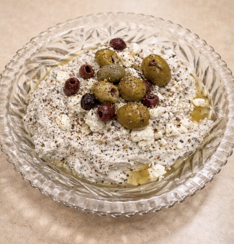 A glass bowl filled with a creamy dip, topped with green and black olives, crumbled feta cheese, cracked black pepper, and a drizzle of olive oil, placed on a beige countertop.