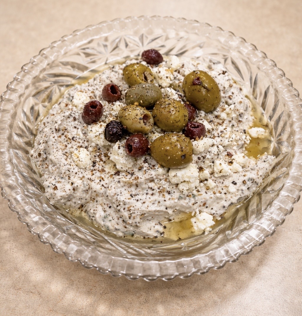 A glass bowl filled with a creamy dip, topped with green and black olives, crumbled feta cheese, cracked black pepper, and a drizzle of olive oil, placed on a beige countertop.